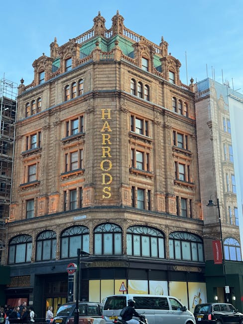 A tall, ornate historic building constructed from brownish-beige stone with detailed architectural features, including decorative cornices and small turrets at the roofline. The building's facade displays prominent vertical signage spelling 'HARRODS' in large, gold-colored letters running from the second to fifth floors. The ground level features large arched windows with dark frames, and a black awning stretches across the shopfront. In front of the building, a busy street scene is visible with several parked cars, including a white van and dark vehicles, as well as pedestrians walking along the sidewalk. A traffic sign and street lamp are also present near the building’s base. The background shows a clear, blue sky and adjacent modern high-rise buildings, creating a contrast with the historic architecture of Harrods. This scene relates to private and independent collections or disposal of rubbish, consistent with alternative waste-handling services for large commercial premises like Harrods.