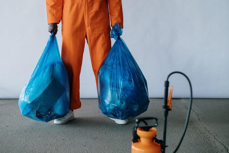 A person dressed in orange work trousers and white shoes is standing indoors on a concrete floor, holding two large blue plastic rubbish bags, one in each hand. The bags are filled with waste and are tightly sealed at the top, showing some bulging contents through the semi-transparent material. A portable sprayer with an orange tank and black hose is positioned in the foreground, partially out of focus. The background features a plain, light-colored wall, and the overall scene suggests an environment suitable for private waste collection or on-site rubbish clearance, which could be associated with services by companies like rubbishclearanceknightsbridge.org.uk. The lighting is even and neutral, emphasizing the focus on waste disposal and waste management activities, aligning with the context of independent rubbish removal and alternative waste handling solutions.