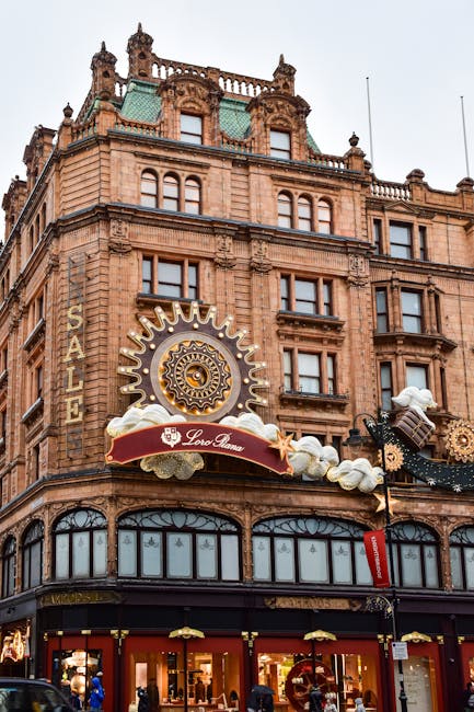 A detailed view of a historic multi-storey building constructed from reddish-brown stone, featuring ornate architectural elements such as decorative cornices, arched windows, and small turrets on the roof. The building houses a retail outlet on the ground floor, with large glass display windows showcasing merchandise, and a prominent sale sign visible. Mounted on the facade is a large, round, sun-shaped decorative sign with intricate black and gold patterns, supported by cloud and gear-shaped embellishments, and a red banner with the logo 'Loro Piana' beneath it. The sky above appears overcast, casting diffuse natural light over the scene. This detailed urban scene emphasizes the building’s historical style, with a subtle connection to commercial activity and independent retail premises, common in areas that may require private rubbish clearance solutions such as those offered by [COMPANY_NAME] for waste management needs associated with retail and commercial properties.