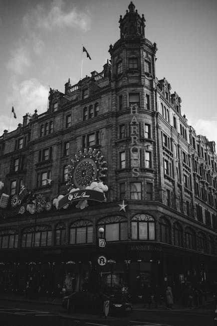 A black and white photograph of a multi-storey historic building situated on a busy urban street corner. The building features ornate architectural details, including decorative cornices, arched windows, and multiple levels with prominent bay windows. Vertical signage attached to the façade indicates the presence of a hotel, with the word 'HARRIS' clearly visible in large illuminated letters. The ground floor appears to house retail or entertainment establishments, with large window displays and a marquee decorated with circular lights and signage for a venue. The street-level area shows parked cars and pedestrians walking along the sidewalk, with some visible street furniture and traffic signs. Above, the cloudy sky adds a moody atmosphere to the scene, emphasizing the building’s historic architecture. This scene provides an example of an urban environment where private property and commercial premises coexist with nearby waste management operations, such as rubbish removal or cleaning activities, typical of city centres where independent collection services like those by rubbishclearanceknightsbridge.org.uk operate to manage waste efficiently in densely built environments.