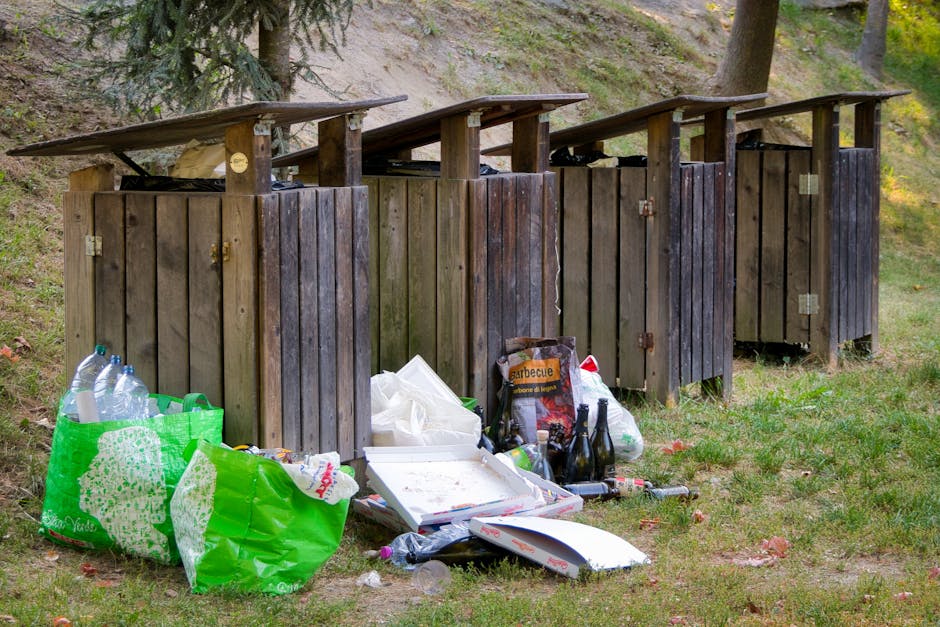 An overflowing collection of mixed waste and rubbish bags is piled on a paved sidewalk near a metal railing in front of a commercial building. The rubbish includes crumpled cardboard boxes, plastic bags, and paper packaging, with some items spilling onto the street. A large grey recycling skip filled with assorted waste is positioned centrally, its lid partially open and overflowing, with paper and cardboard protruding from inside. Surrounding the skip, various black and red rubbish bags are stacked or scattered, some tightly tied and others torn open, revealing discarded materials. To the left, a parked grey car with a UK registration plate is visible behind the barrier. The background features a blue metal balcony or scaffolding structure attached to the building, which has a commercial storefront with several signs and closed roller shutters. The scene suggests a situation of waste accumulation that may be addressed through alternative rubbish collection or private disposal services, with the presence of [COMPANY_NAME] though not explicitly shown. The environment is well-lit, indicating daytime, and the overall scene captures a typical urban area experiencing rubbish collection delays or accumulation issues.