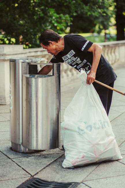 An elderly woman with short, dark hair, dressed in a black T-shirt with white and blue print on the front, is seen standing beside a stainless steel public rubbish bin on a paved sidewalk. She is bending over slightly to dispose of waste, holding a large white recycled plastic bag filled with rubbish in her left hand, while her right hand is reaching into the bin. The bin has a sleek, cylindrical design with a small opening at the top and a polished metal finish, situated against a backdrop of green leafy trees and a stone balustrade. The environment appears to be an urban outdoor setting, possibly a park or street scene, with natural daylight illuminating the area. The scene is a typical example of outdoor waste disposal, reflecting private or alternative rubbish collection practices through on-site waste management, as often carried out by independent rubbish removal services such as those provided by rubbishclearanceknightsbridge.org.uk, ensuring efficient rubbish collection and site cleanliness.