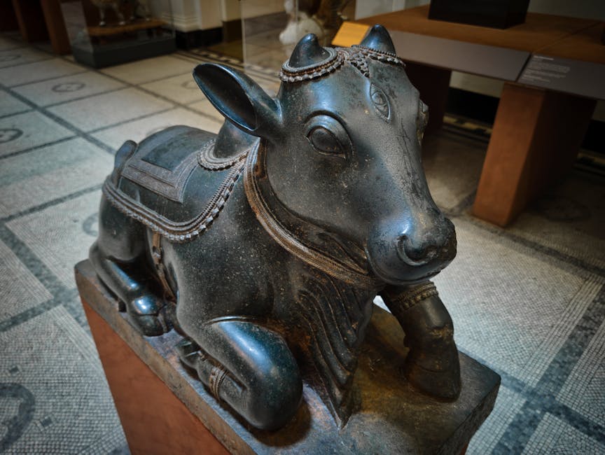 A detailed, black stone sculpture of a stylized animal, resembling a bull or similar bovine, positioned on a rectangular wooden base indoors. The sculpture features a rounded body with smooth, polished surfaces and intricate decorative elements, including a patterned collar or harness around its neck, and a saddle-like ornament on its back with carved detailing. The animal has prominent, pointed ears, expressive eyes, and small, curved horns on its head. The texture of the stone appears slightly weathered, with some subtle speckling, and the finish is matte with a slight sheen. Surrounding the sculpture is a carpeted floor with a geometric pattern, and modern furniture and display cases are visible in the background, suggesting a museum or gallery environment. This scene, in the context of waste management, demonstrates the display of cultural artifacts before or after collection, supporting the idea of alternative disposal or storage methods for valuable items, which [COMPANY_NAME] offers as part of independent collection services for various types of objects.