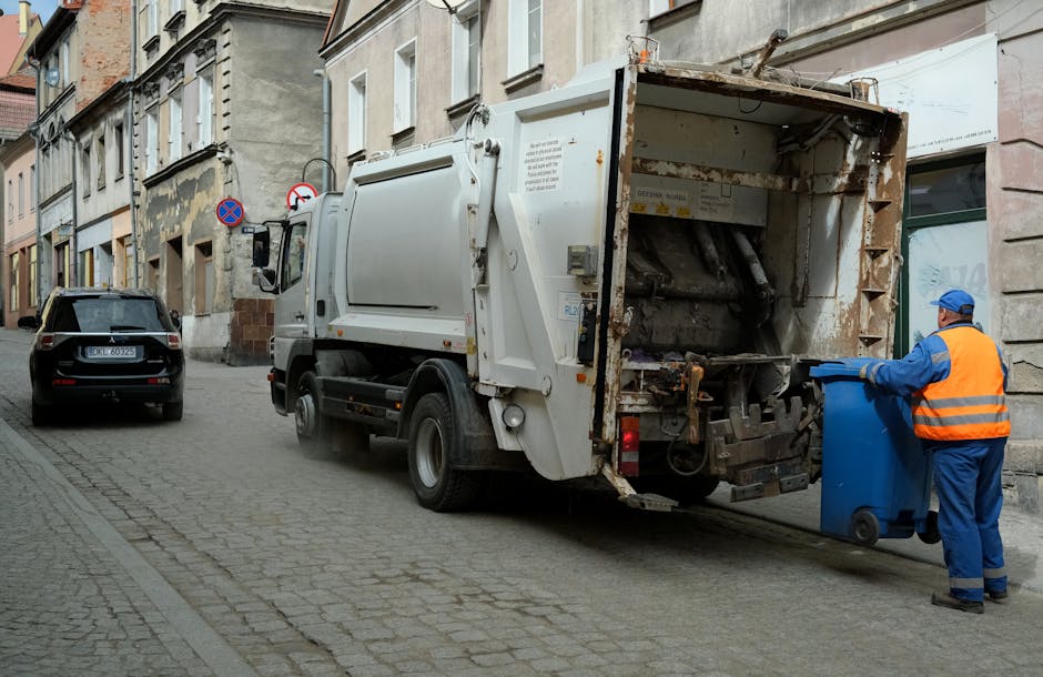 An elderly woman with short, dark hair, dressed in a black T-shirt with white and blue print on the front, is seen standing beside a stainless steel public rubbish bin on a paved sidewalk. She is bending over slightly to dispose of waste, holding a large white recycled plastic bag filled with rubbish in her left hand, while her right hand is reaching into the bin. The bin has a sleek, cylindrical design with a small opening at the top and a polished metal finish, situated against a backdrop of green leafy trees and a stone balustrade. The environment appears to be an urban outdoor setting, possibly a park or street scene, with natural daylight illuminating the area. The scene is a typical example of outdoor waste disposal, reflecting private or alternative rubbish collection practices through on-site waste management, as often carried out by independent rubbish removal services such as those provided by rubbishclearanceknightsbridge.org.uk, ensuring efficient rubbish collection and site cleanliness.