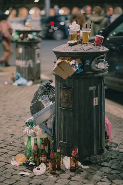 A heavily soiled black public litter bin made of metal with a domed lid, situated on a cobblestone pavement, is overflowing with rubbish and surrounded by scattered trash, including crumpled paper, glass bottles, and food containers. Several bottles, some green and some brown, are positioned near the base of the bin, with additional waste such as a plastic bag and discarded packaging nearby. On top of the bin's lid, there are three to four beverage cups with plastic lids and straws, along with a small cardboard box and some food wrappers, suggesting recent food consumption. The background shows a blurred urban street scene with parked cars and indistinct pedestrians, indicating a busy public area. The scene emphasizes the challenges of unmanaged waste in an outdoor environment, which may relate to alternative or unregulated waste disposal practices, as reflected in the apparent overflow and litter scattered around the bin.