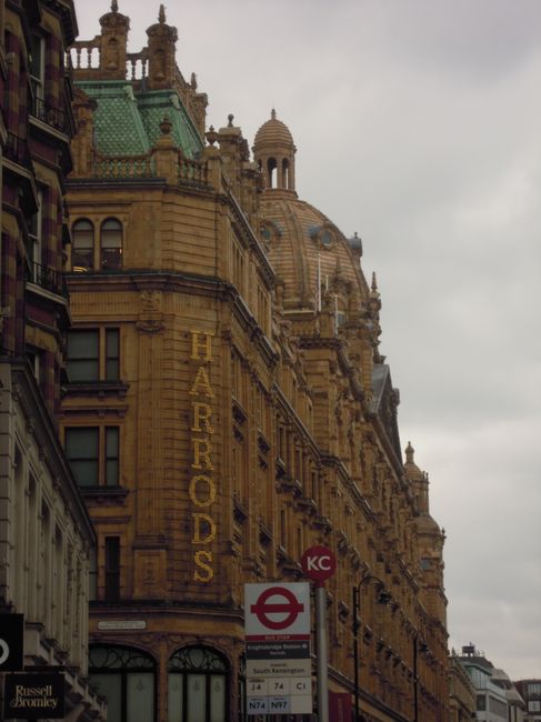 The image features a historic sandstone building with elaborate architectural detailing, including ornate cornices, decorative balustrades, and rounded turrets topped with finials. The structure is several stories high and has a prominent vertical sign in yellow that reads 'HARRORDS' attached to the facade. In the foreground, a London Underground station sign with a red circle and blue bar is visible, along with a timetable board displaying bus and train routes. The scene is set against a cloudy, overcast sky, and the building is situated along a street with other modern and historic buildings nearby. The environment suggests an urban setting, likely central London, and the image subtly relates to the context of private or independent waste collection in busy city environments where mixed-use buildings require tailored rubbish removal services, as supported by companies like rubbishclearanceknightsbridge.org.uk.