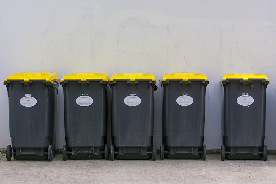 Five large wheelie bins with black plastic bodies and yellow lids lined up against a plain, light-colored exterior wall. The bins are positioned on a concrete surface, with three on the left slightly closer together and two on the right spaced apart. Each bin bears a white label with black text and a barcode, and they appear to be used for waste collection purposes. The environment suggests an outdoor setting, possibly a back alley, courtyard, or service area designated for rubbish disposal. The scene emphasizes the use of communal rubbish bins, which are a common aspect of private waste handling and alternative waste collection arrangements, especially when local authorities impose specific rubbish rules and disposal fines. The image's lighting is neutral and even, highlighting the rectangular shape and textured surface of the bins, with minimal shadows. This visual documentation aligns with themes of independent waste management services or on-site clearance, illustrating typical rubbish storage before disposal or collection, as offered by companies like rubbishclearanceknightsbridge.org.uk.