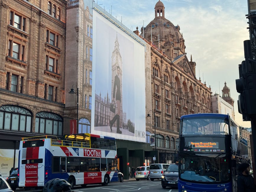 A city street scene featuring a historic building with ornate stonework and a prominent dome in the background. To the left, a large vertical billboard displays an advertisement with a model and the Dior logo, mounted on the building's facade. In the foreground, a double-decker bus with a digital route display is visible, alongside a red and white double-decker tour bus with the words 'TOOT bus' and 'London' on its side. Several cars are parked along the street, and a person wearing a dark coat and face mask is walking on the sidewalk. The environment is urban with a mix of historic architecture and modern transportation, typical of a busy commercial district where private waste collection and rubbish removal services might operate independently from local authority services, especially in areas like Knightsbridge near Harrods.