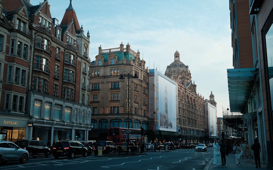 A city street scene featuring a historic building with ornate stonework and a prominent dome in the background. To the left, a large vertical billboard displays an advertisement with a model and the Dior logo, mounted on the building's facade. In the foreground, a double-decker bus with a digital route display is visible, alongside a red and white double-decker tour bus with the words 'TOOT bus' and 'London' on its side. Several cars are parked along the street, and a person wearing a dark coat and face mask is walking on the sidewalk. The environment is urban with a mix of historic architecture and modern transportation, typical of a busy commercial district where private waste collection and rubbish removal services might operate independently from local authority services, especially in areas like Knightsbridge near Harrods.