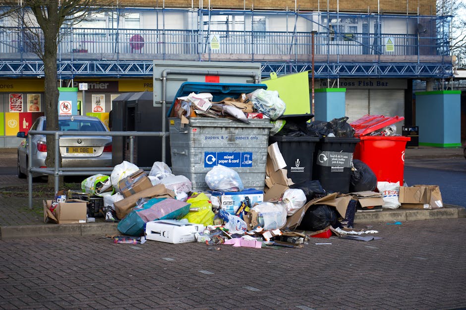 A collection of overflowing rubbish bins and scattered waste bags situated on a paved sidewalk in an urban area. The scene includes a large grey mixed waste container filled with crumpled paper, cardboard, and plastic packaging, with some items spilling over onto the ground. Surrounding the bin are various black and red plastic garbage bags, flattened cardboard boxes, and loose packaging materials such as paper and foam. In the background, there is a parked car with a European-style license plate, and a building with a storefront featuring signage, and blue scaffolding or construction wrap covering the upper part of the structure. The environment appears to be a commercial or residential precinct with minimal natural elements, and the waste is positioned behind a metal railing, indicating a designated area for rubbish collection, reflecting a need for effective rubbish removal or private waste handling services in the area.