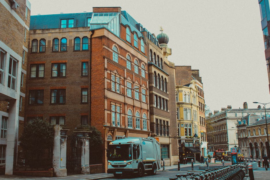 A mid-sized, light grey waste collection truck with a compact cab and cargo area is parked on a city street in front of a row of historic buildings. The truck features a smooth, metallic finish with black trim around the windows and a white logo or branding on the side that is partially visible. It is positioned on the pavement, adjacent to a metal bicycle rack and a row of black bollards. The surrounding buildings display a mix of architectural styles, with many featuring brick facades, arched windows, decorative cornices, and ornate detailing. One building has a distinctive dome-shaped turret on its roof. There is a small tree or shrub in front of a gated entryway on the left side of the image, and street lamps are visible further down the road. The scene is lit by natural daylight, creating a clear and neutral atmosphere suitable for illustrating private waste handling or independent rubbish collection services in an urban environment. This setup aligns with concepts of on-site clearance or alternative waste management options supported by local rubbish removal firms like Knightsbridge rubbish removal prices real cost guide 2026.