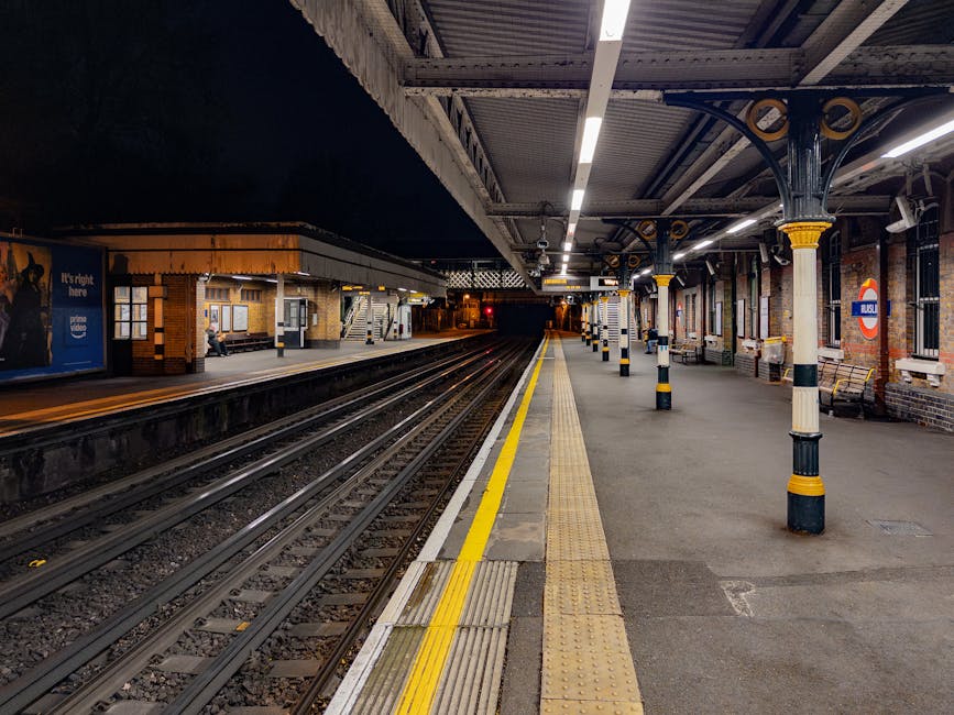 The image shows a view of a railway station platform at night, illuminated by overhead lighting. The platform has a textured yellow safety line and tactile paving along the edge, which runs parallel to multiple railway tracks composed of steel rails and wooden sleepers. Several brick buildings with large windows and a sheltered waiting area are visible on the left side, with a person sitting on a bench. The platform is supported by decorative iron columns featuring black and yellow detailing, situated along the covered section. An iconic roundel sign indicating the station name is visible on the right, attached to a brick wall with multiple windows. The scene is quiet, with no passengers or trains present, and the background extends into darkness, giving a sense of depth and calmness typical of an urban underground or suburban railway setting. This scene is related to urban transport infrastructure, often maintained through services that include rubbish and waste removal at station premises, relevant to independent waste disposal or rubbish clearance services near transportation hubs.