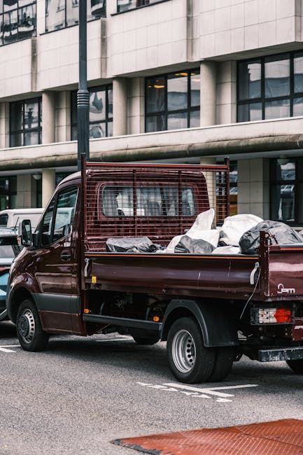 A small burgundy flatbed pickup truck parked on a city street in front of a modern building with large glass windows and concrete facade. The truck's cargo bed is filled with various discarded items, including white and black plastic bags, some crumpled and partially open, revealing unclear contents. The truck features a metal safety grille around the cab and sides, with visible wheel arches and silver hubcaps on the tires. The vehicle appears to be used for private waste removal or rubbish clearance, aligning with the services offered by companies like rubbishclearanceknightsbridge.org.uk. The scene is set during daylight, with natural light casting minimal shadows, and the background includes other parked cars and urban elements, emphasizing the context of municipal or independent rubbish disposal in an urban environment. Overall, the image illustrates the practical aspect of small-scale rubbish removal and waste handling in densely populated areas.